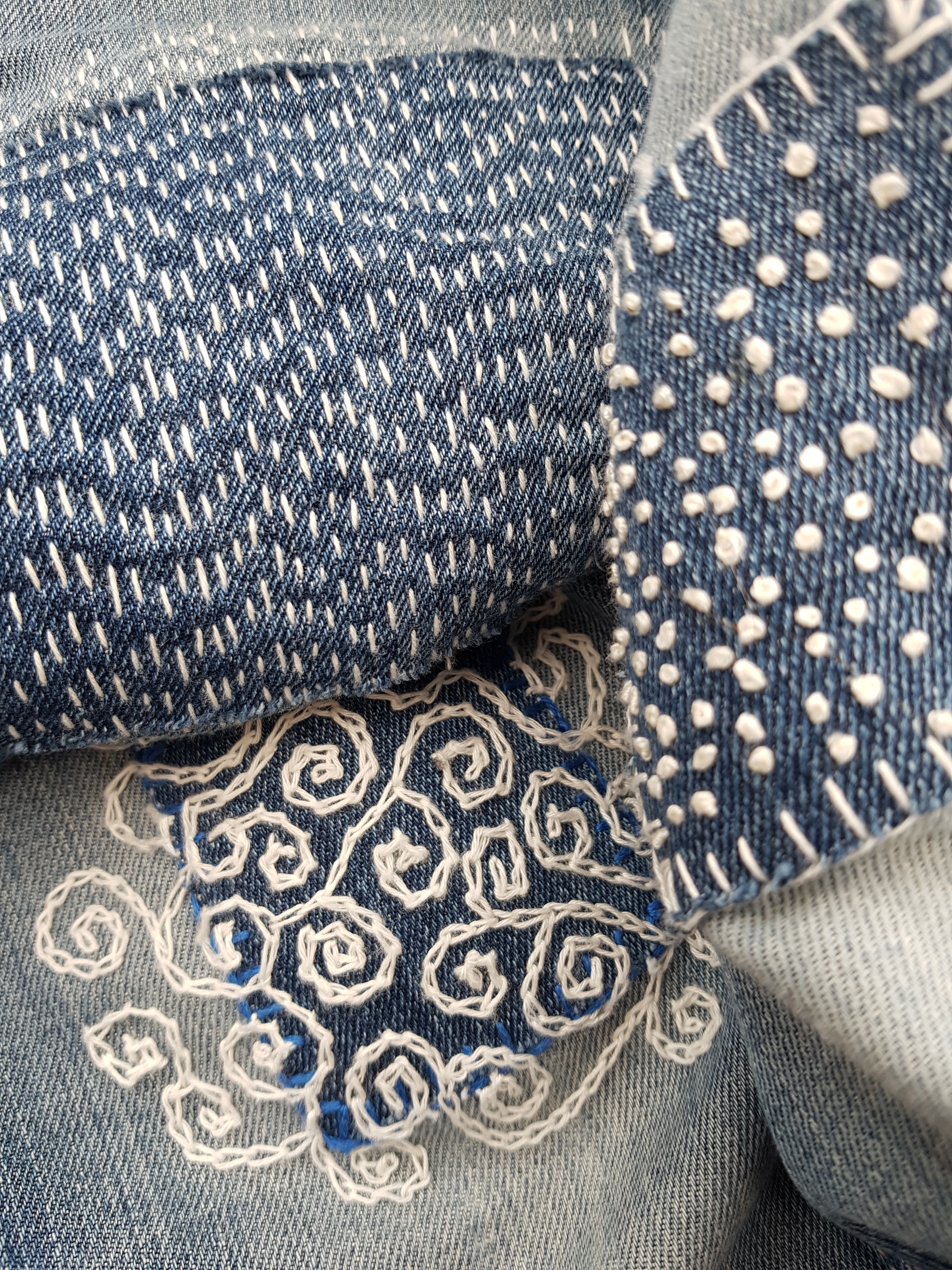 Close-up photo of the knees of a pair of jeans which have had decoratively stitched (boro, French knots, chain stitch) denim patches applied to not just fix but enhance the holes