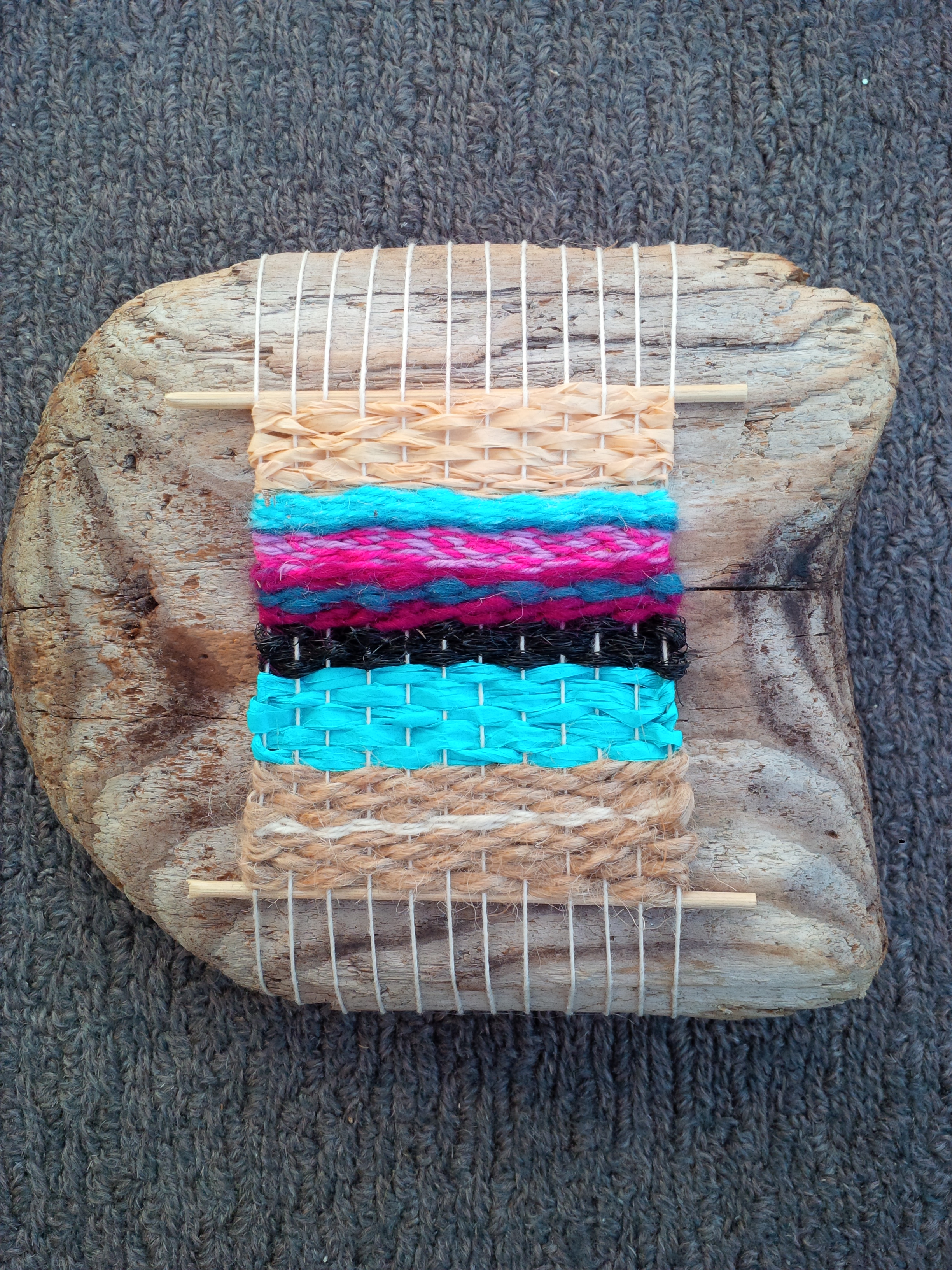 A chunk of silvery weathered driftwood, with a warp of threads and weaving onto the warp in raffia and yarns, in turquoise, pinks, beige and black